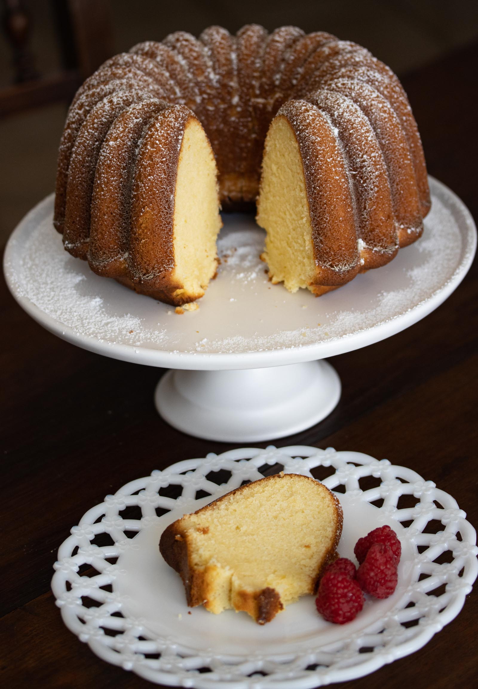 Lemon Pound Cake on a white cake stand and a slice served on a white dish with raspberries