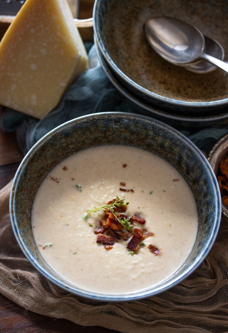 Roasted Cauliflower Soup in a blue bowl with bacon bits and parmesan cheese next to it