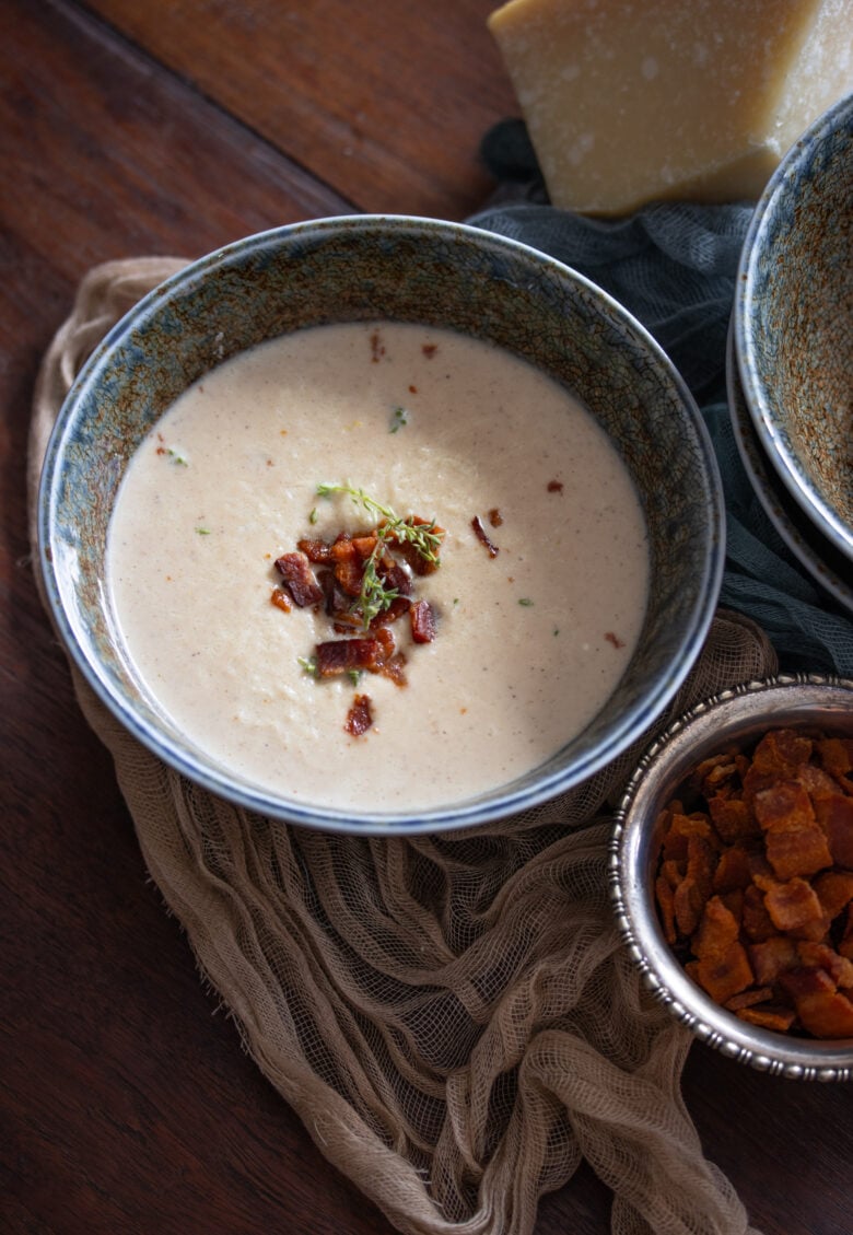 Roasted Cauliflower Soup in a blue bowl with bacon bits and parmesan cheese next to it