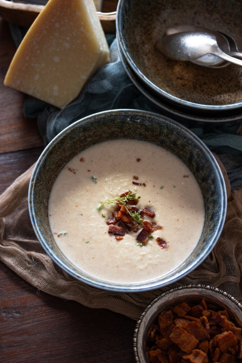 Roasted Cauliflower Soup in a blue bowl with bacon bits and parmesan cheese next to it