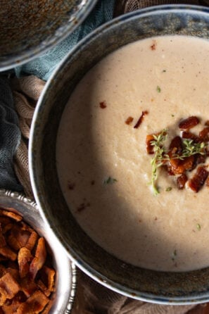 Roasted Cauliflower Soup in a blue bowl with bacon bits and parmesan cheese next to it